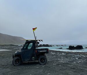 Polaris side by side vehilce with board on top and surf life saving flag in back traybody, parked on black sand beach