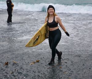 Woman wearing leggings, sports bra and beanie walks from water holding surf board