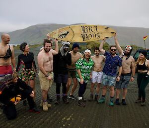 Group of expeditioners dressed ready for a swim gather and hold board aloft above heads, smiling to camera