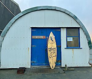 Wooden surf board resting against a building which is a long domed building with blue front door