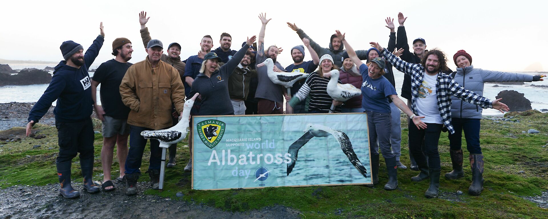 A group of expeditioners standing behind a banner reading "world albatross day" and some are holding albatross decoys, others are spreading their arms like wings