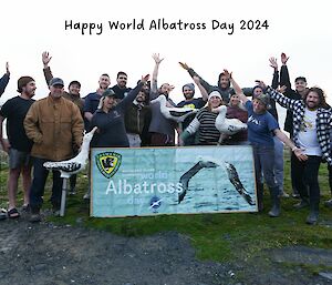 A group of expeditioners standing behind a banner reading "world albatross day" and some are holding albatross decoys, others are spreading their arms like wings