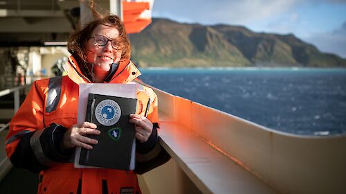 A woman in orange hi vis stands on a ship's deck with an island in the background.
