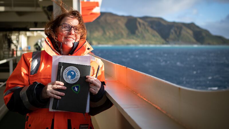 A woman in orange hi vis stands on a ship's deck with an island in the background.