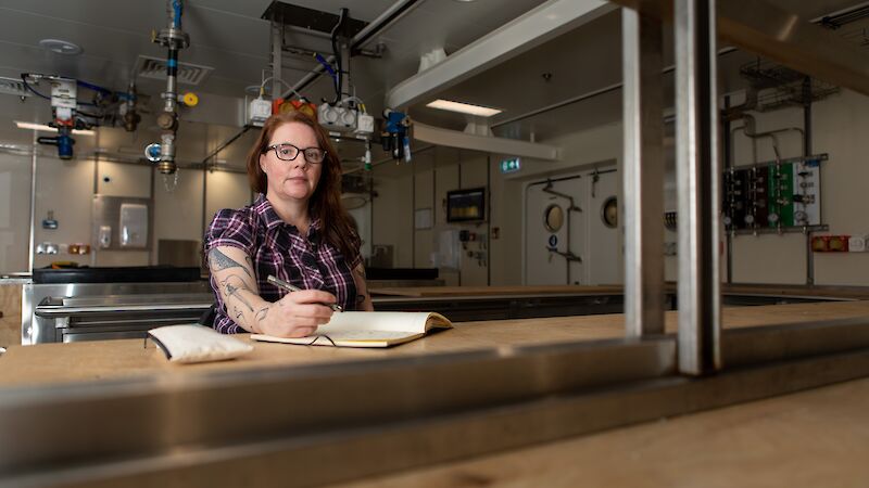 A red-headed woman in a check shirt sits in a science lab with a notepad.