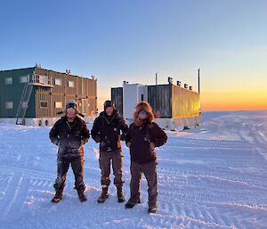 Three people in outdoor gear stand outside a shed at dawn or dusk