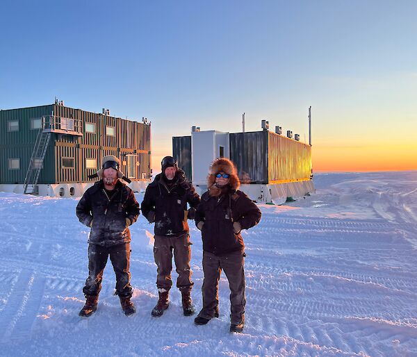 Three people in outdoor gear stand outside a shed at dawn or dusk