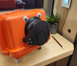 An orange camera with googly eyes stuck on, sitting on a table