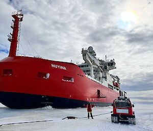 A big red ship is sitting surrounded by ice with a person and a tractor in front