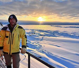 A man in yellow stands in front of a hut with the sun rising over a snowy landscape