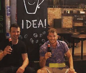 Two men sitting on stools holding beers and smiling