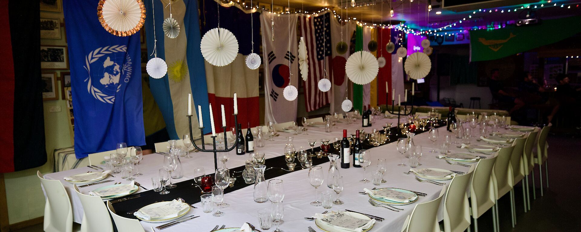 Large table seating 24, with white tablecloth, candles down centre, formal settings at each plate. Paper fans, fairy lights and flags hanging over and aroudn the table.