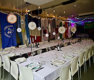 Large table seating 24, with white tablecloth, candles down centre, formal settings at each plate. Paper fans, fairy lights and flags hanging over and aroudn the table.
