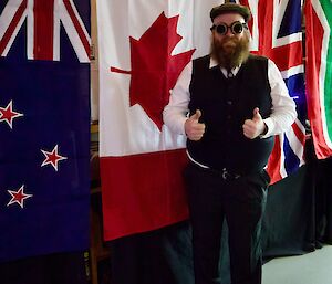 Man stands in front of Canadian flag, wearing waistcoat and steampunk goggles and peaked cap