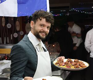 Man in suit, wearing white apron, carries two platters of canapes, and smiles to camera