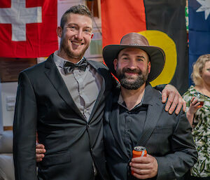 Two men in formal attire, one with arm over shoulders of the other who is wearing a hat, smile to the camera