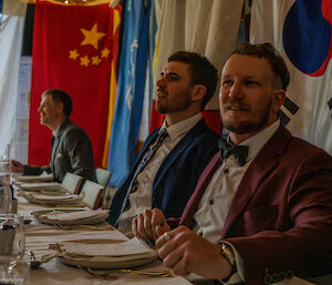 Three men sitting at the dinner table, wearing formal attire, sit and listen to speeches