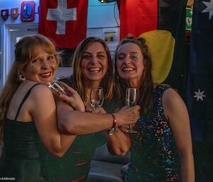 Three women link arms while holding champagne glasses, and smile to camera
