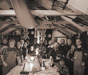 Group photo of whole station inside old wooden A-frame hut, sepia colours, people wearing older style expeditioner clothing including overalls, chequered shirts and woollen jumpers, flags lining the walls