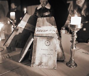 Sepia photo of pyramid-shaped wooden box labelled “Mawson 1916” on a table with two old station log books, next to a candle