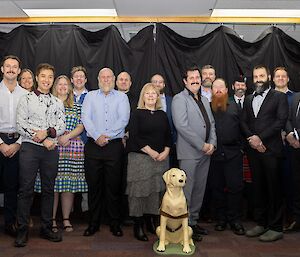 Group photo of whole station dressed in formal attire in front of a black curtain. Guide dog Stay 2.0 in the foreground