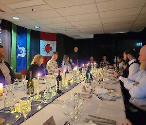 People sitting around a long dining table, with a white tablecloth and blue table runner, decorated with candles and fairy lights. People in formal attire. Flags in the background