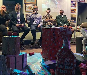 A table of colourfully wrapped gifts in the foreground, people sitting on stools in a semi-circle in the background