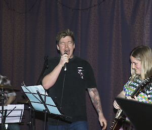 Photo of three members of the band in from of a purple curtain background. Rhys is on drums on the left, Braiden is belting out lyrics in the centre with his eyes closed and mouth wide, and Sophie is playing guitar on the right