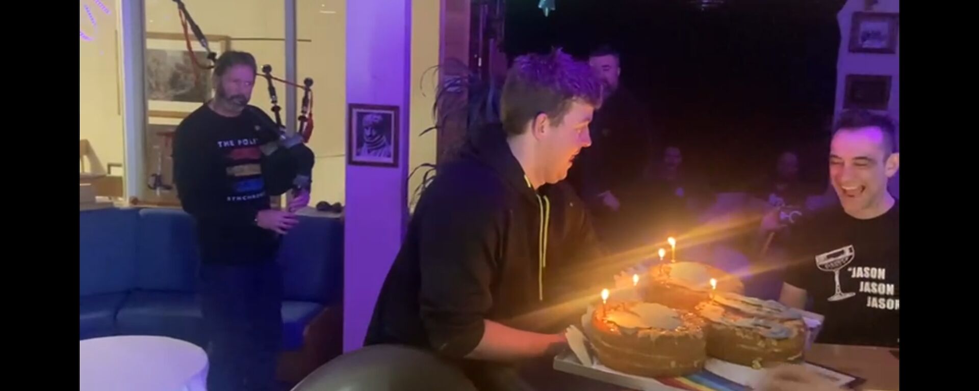 Peter standing to the left playing bagpipes, while Braiden presents a tray holding three round cakes with candles sitting on a rainbow to Jason wearing a big smile on the right
