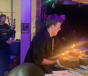 Peter standing to the left playing bagpipes, while Braiden presents a tray holding three round cakes with candles sitting on a rainbow to Jason wearing a big smile on the right