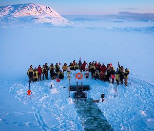 the Casey station expeditioners gathered around the cut out ice hole as the sun rises with Budnick hill in the background