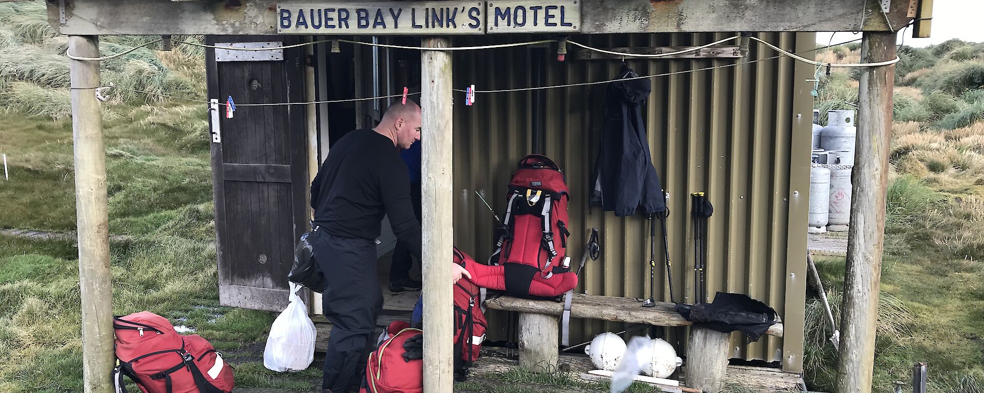 Looking front on at small corrogated iron covered hut with verandah at front, man and four red hiking packs on verandah