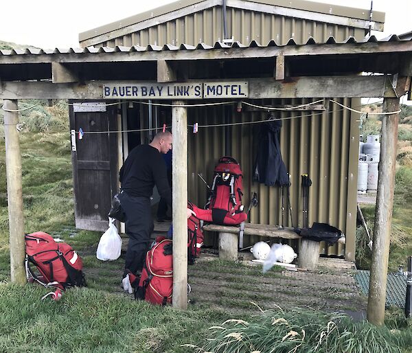 Looking front on at small corrogated iron covered hut with verandah at front, man and four red hiking packs on verandah