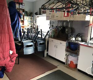 Inside of hut, showing kitchen bench with gas hotplate and sink, and four seater table with chairs upside down on table, in foreground coathangers and sleeping bag hanging from ceiling