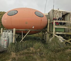 Fiberglass orange hut, shaped like a smartie, with wooden structure as entry porch on right side, gas bottles on left side