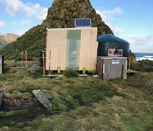 Small wooden hut beside coverted water tank hut, in the background a small grass covered peak and then behind the coastline