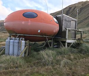 Fiberglass orange hut, shaped like a smartie, with wooden structure as entry porch on right side, gas bottles on left side