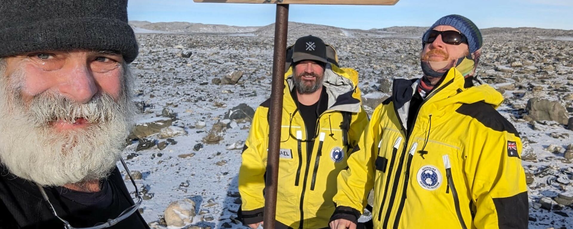 two men in Antarctic yellow suits and a man with a big beard stand near a signpost
