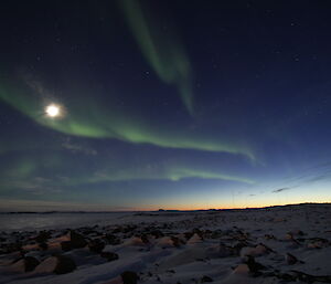 Ribbons of green aurora streak across a navy blue sky towards an soft orange glow on the horizon, illuminating snow-covered rocks below.