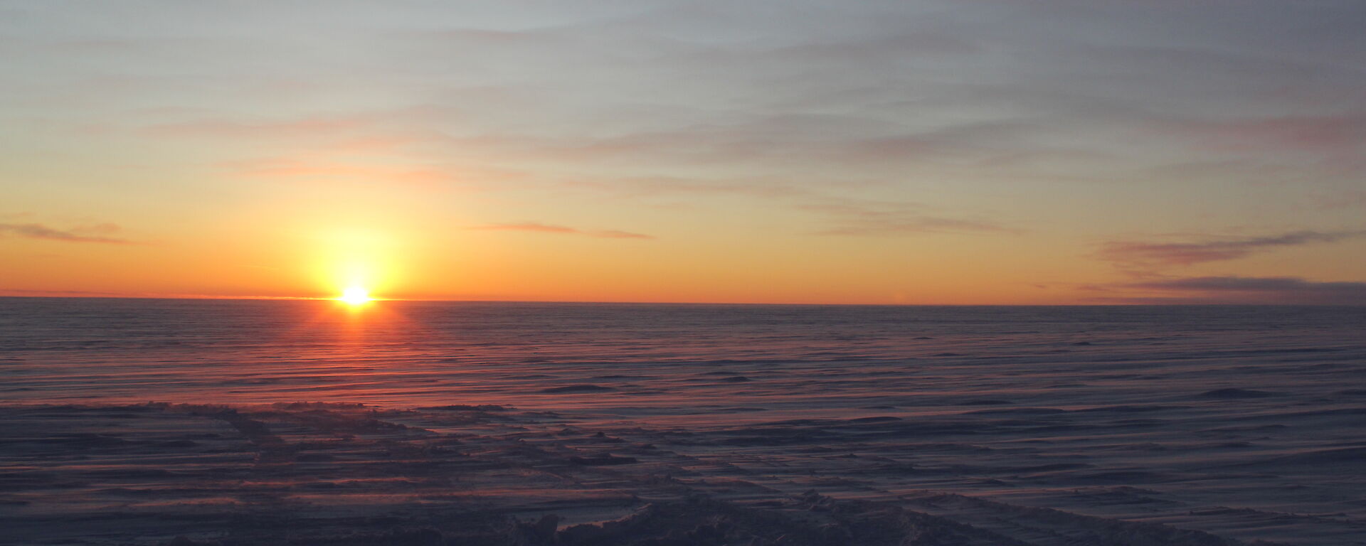 The sun, low on the horizon, casts a rosy glow across a flat, snow-covered landmass.