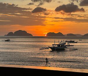 the sun sets deep in the background of the coastal landscape behind an island, ashore a silhouette person swims