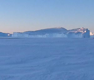 A group of emperor penguins at the left of the panoramic photo on a light blue ice plain. In the distance are many huge icebergs and a large huddled group of hundreds of emperor penguins.