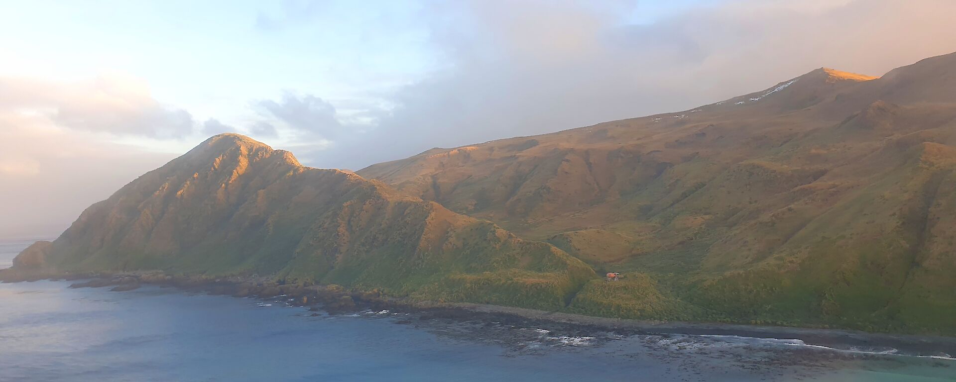 Looking across the sea from an aircraft at a mountain range with small hut on a ridge line about a third of the way up