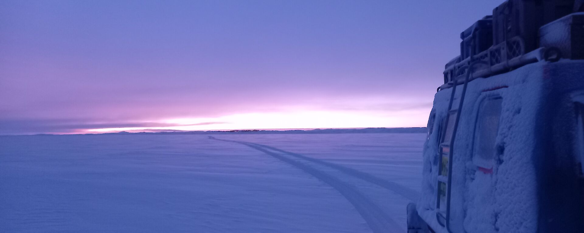 A tractor with tractor tracks behind it in the snow, under a pale sunrise or sunset