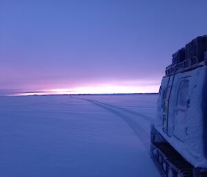 A tractor with tractor tracks behind it in the snow, under a pale sunrise or sunset