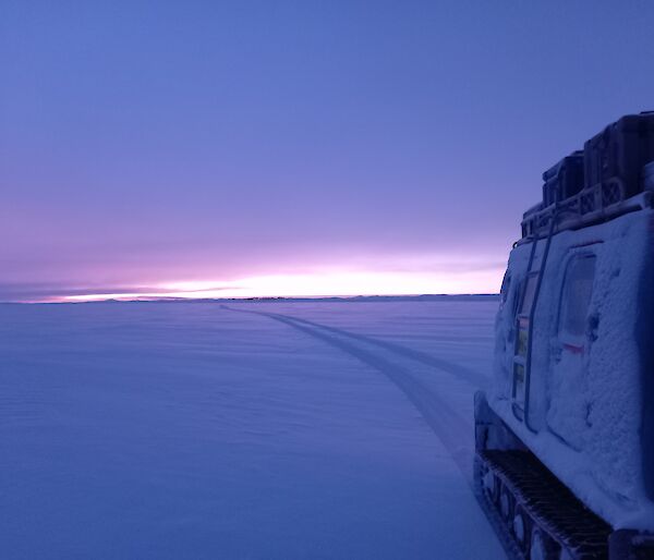 A tractor with tractor tracks behind it in the snow, under a pale sunrise or sunset