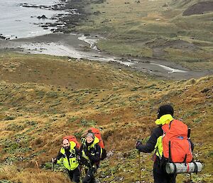 Three walkers heading up a steep hill with a rocky shoreline in the background