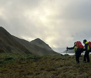 Two walkers withpoles head across a green landscape with mountains to the left and station buildings ahead