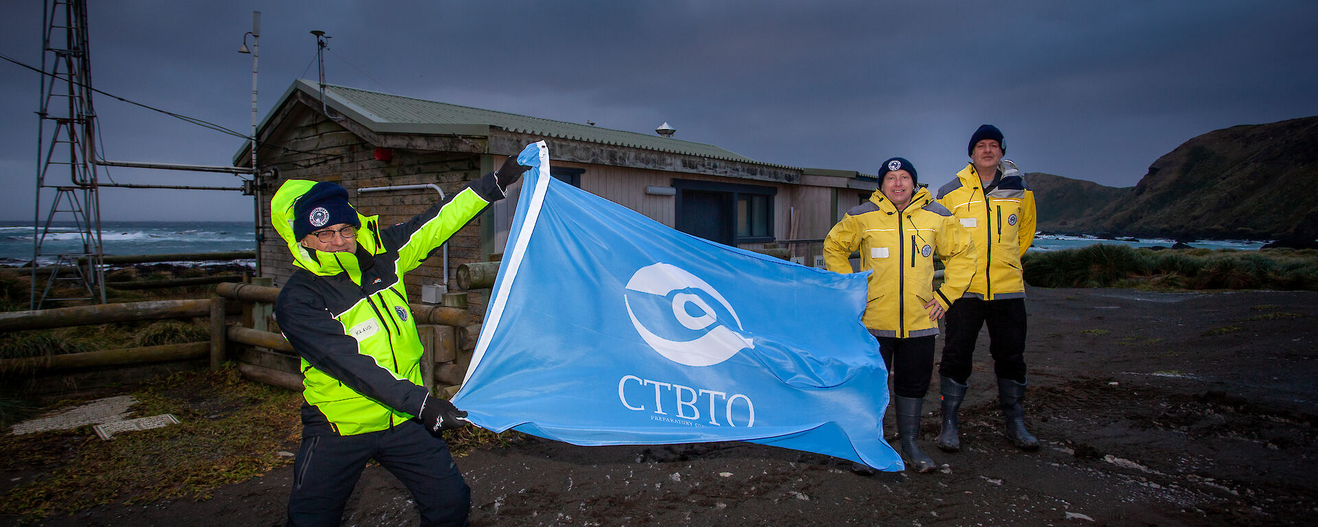 A man in high vis holds a CTBTO flag out in the wind with a shed behind him, and two colleagues in yellow to the right.