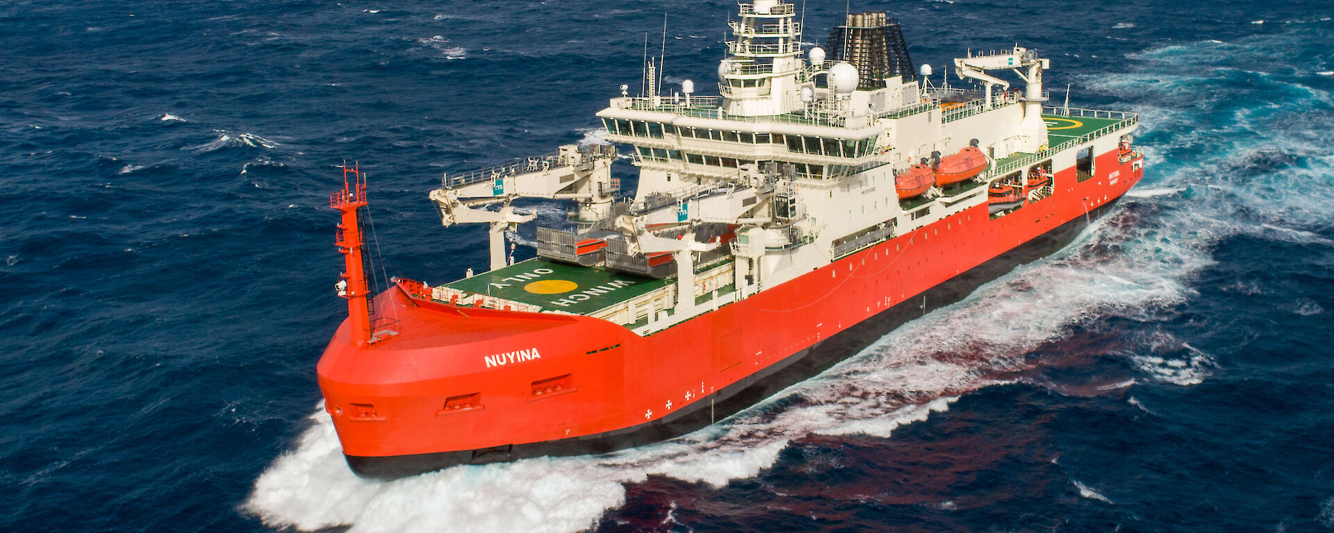 An aerial drone photo of an orange and white icebreaker ship sailing though a blue ocean